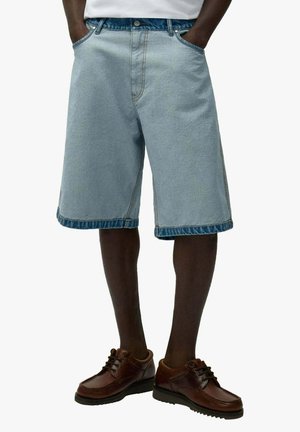 Uomo che indossa pantaloncini di jeans azzurri fino al ginocchio con orli risvoltati e scarpe stringate in pelle marrone, in piedi con le mani in tasca.