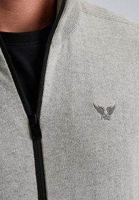 Grau farbener Zip-Kapuzenpullover aus weichem Material mit geripptem Kragen. Verfügt über ein Logo mit Flügeln auf der linken Brust und eine schwarze Reißverschlussschnalle.