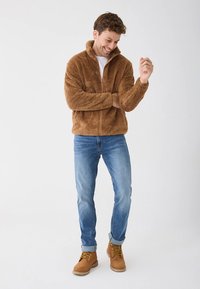 Ruda pūkuota striukė su užtrauktuku ir aukšta apykakle, derinama su mėlynais džinsais ir šviesios rudos spalvos žygiavimo batais. Audinys turi minkštą, tekstūruotą išvaizdą.