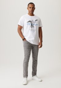 Biała bawełniana koszulka z graficznym nadrukiem przedstawiającym anioła oraz niebieskim tekstem, zestawiona z jasnoszarymi dżinsami i białymi trampkami.