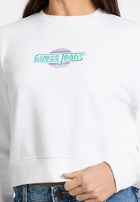 Valkoinen lyhyt cropped-huppari, jossa on teal- ja purppuravärinen kirjailtu "Guess Jeans" -logo, tekstin takana on pyöreä purppuraväri.