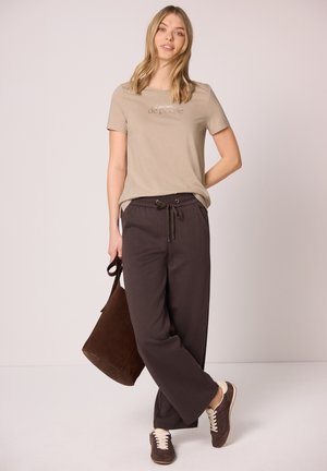 Žena v béžovém krátkém rukávu s nápisem, volné tmavé kalhoty, hnědé tenisky, držící velkou tmavě hnědou tašku na jednobarevném pozadí.