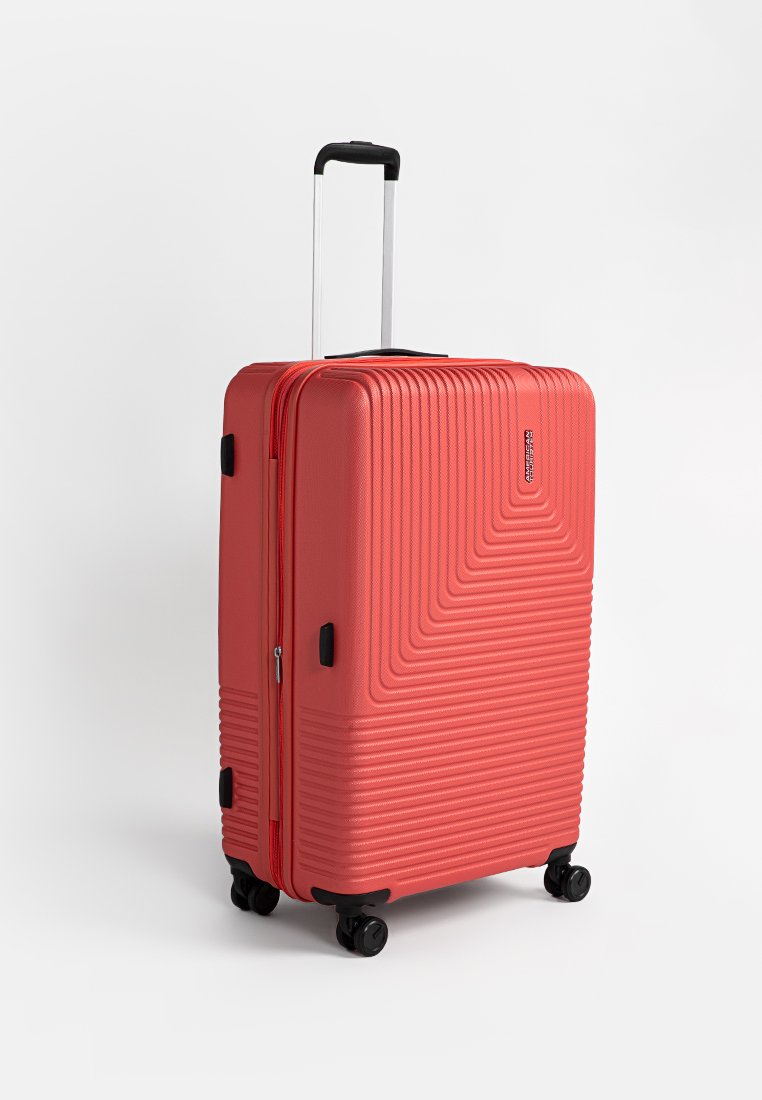 Valise rigide en corail avec motif en zigzag texturé, une poignée télescopique et quatre roues noires pour la mobilité. Fermeture à glissière robuste.