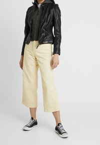 Musta nahkamoottoripyörätakki, harmaa huppari alla, keltaiset leveälahkeiset housut ja mustat lenkkitossut valkoisilla pohjilla. Yhteensopiva minimalistinen muotoilu.