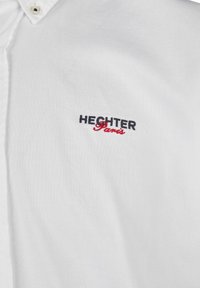 Witte, kortge mouwen shirt van katoen. Kenmerkt zich door een kraag met knopen en geborduurd "HECHTER" in marineblauw en "Paris" in rood op de borst.