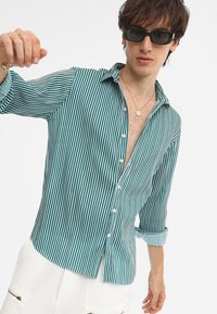 Grün und weiß gestreifte, lässig geschnittene Hemdbluse mit langen, hochgerollten Ärmeln und einem Kragen, der einen Teil der Brust freilegt.