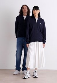 Maglione zip-up navy, gonna plissettata bianca e sneaker sportive; stile casual con texture variegate; due modelli in piedi contro uno sfondo semplice.