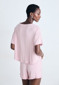 Kvinna med kort lockigt hår som bär en lös rosa topp med korta ärmar och rosa shorts med prickar och volanger i kanten, stående framför en enfärgad bakgrund.
