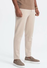 Béžové kalhoty na míru s texturovanou tkaninou, slim fit a čistými liniemi, doplněné šedými slip-on botami a hnědým pleteným topem.