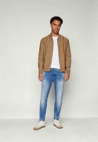 Ung man i beige bomberjacka, vit t-shirt, ljusblå jeans och vita sneakers som står mot en ljus enfärgad bakgrund.