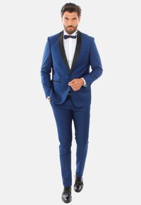 Tuxedo blu con risvolti in satin nero, design aderente, accompagnato da una camicia bianca e un papillon nero; scarpe lucide nere.