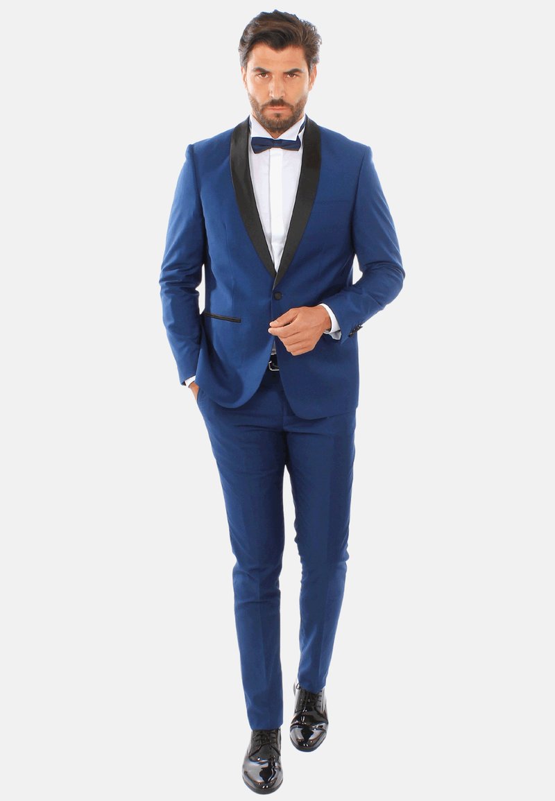 Tuxedo blu con risvolti in satin nero, design aderente, accompagnato da una camicia bianca e un papillon nero; scarpe lucide nere.