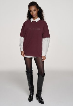 Vrouw met krullend haar die een oversized wijnrode T-shirt draagt over een wit shirt met lange mouwen, doorschijnende zwarte panty's en zwarte kniehoge laarzen.