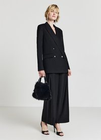 Blazer doppiopetto nero con righe sottili argentate, abbinato a pantaloni a gamba larga. Il modello tiene in mano una borsa in pelliccia nera ed indossa sandali neri con tacco.