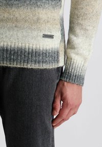Strickpullover mit einem Verlauf aus Creme-, Grau- und Dunkelgrautönen, mit gerippten Bündchen und Saum, begleitet von einem metallischen Logo-Tag.