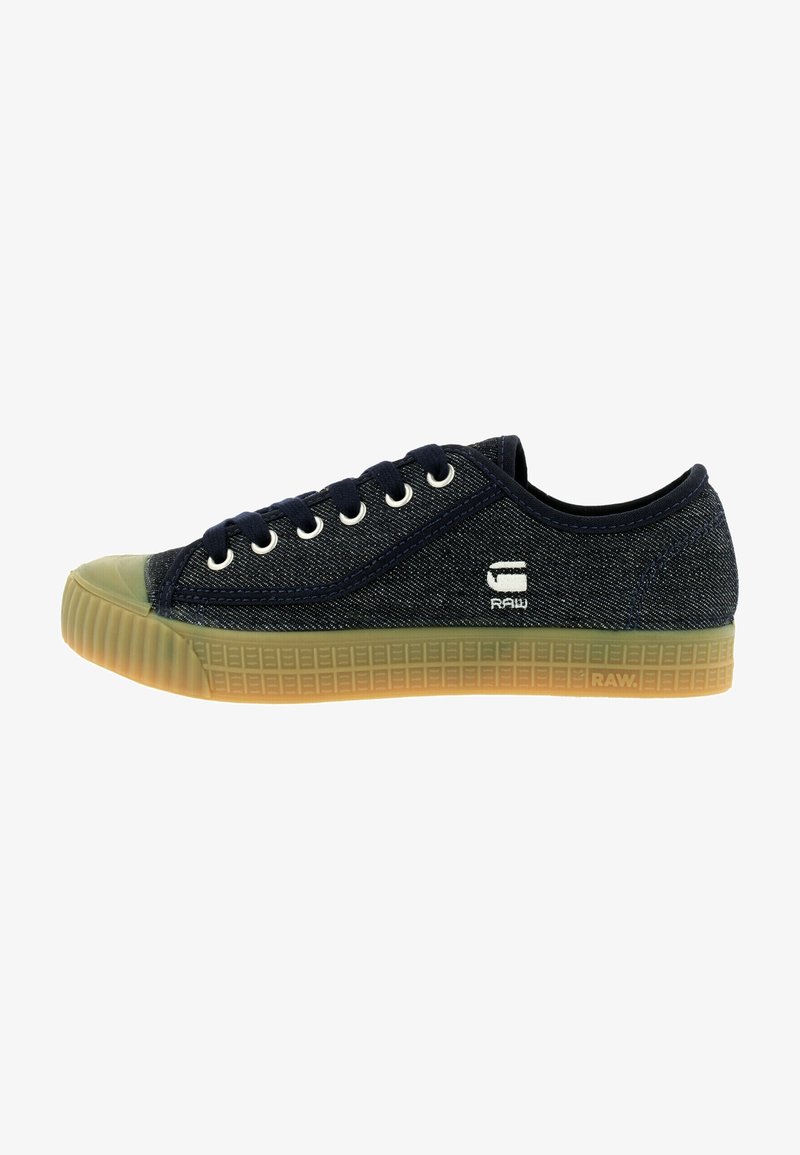 Dark blue canvas sneaker with round toe, rubber sole, white eyelets, and side logo detail. Features textured fabric and contrasting green accents.