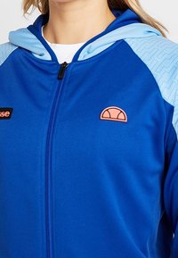 Blå zip-up hoodie med en ljusblå huva, gummiserig rosa logotyp och texturerat blått mönster på axlarna. Mjuk, slät tyg.