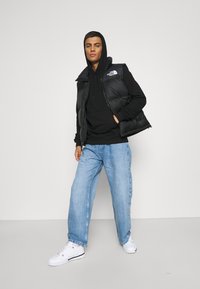 Black padded vest over a black hoodie, paired with loose blue jeans and white sneakers. The vest features a logo on the chest.