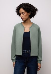 Lysegrøn sweatshirt med lynlås foran, ribbede ærmegab og krave, lavet af blødt stof; båret over en marineblå top og kombineret med mørke denimjeans.