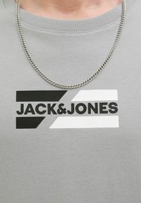 Pilkos spalvos medvilninė marškinė su juodai balta logo, kuriame yra geometrinės formos ir tekstas "JACK&JONES", derinamos su sidabriniu grandininiu kaklo papuošalu.