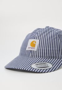 Navy and white striped cap made of fabric with a rounded peak, featuring a yellow logo patch and an adjustable strap label.