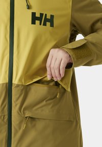 Wasserabweisende Jacke in lebhaftem Gelb-Grün, ausgestattet mit einer Reißverschlusstasche, Logo-Detail und Klettverschluss-Elementen an den Ärmeln. Glattes, strukturiertes Material.
