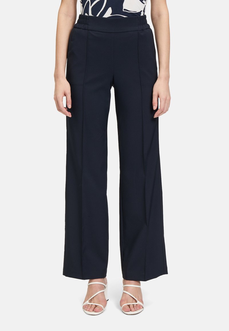 Pantaloni a gamba larga blu navy con una cintura elastica e allungabile. Presentano cuciture frontali e una texture del tessuto liscia. Abbinati a sandali bianchi con cinturini.