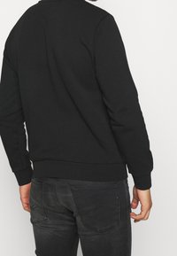 Sudadera negra de manga larga, con puños y dobladillo acanalados, textura suave y un diseño simple, sin adornos. La vista muestra la parte trasera de la prenda.