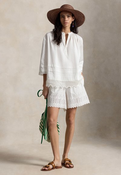 Γυναίκα που φοράει λευκό μπλουζάκι από δαντέλα με μικρές τρύπες και σορτς, καφέ πλεγμένο καπέλο, καφέ σανδάλια, κρατώντας πράσινη και λευκή ριγέ τσάντα.