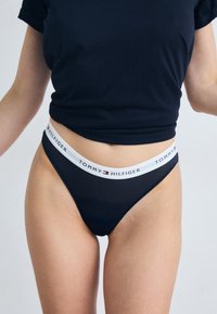 Jūrinio stiliaus juostuota thong tipo apatinė trinkelė su balta ženkline juosta, ant kurios yra „TOMMY HILFIGER“ tekstas. Lygus audinys, minimalios aprėpties dizainas.