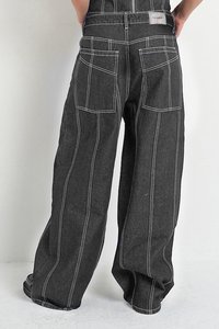 Asmuo vilkintis plačias juodas kelnes su baltomis kontrastingomis siūlėmis, dviem galinėmis kišenėmis ir juodais platforminiais bateliais ant baltų grindų.