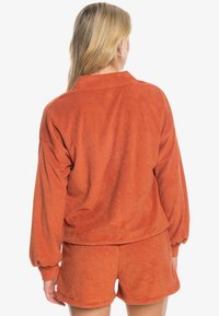 Pomarańczowa bluza polarowa o luźnym kroju, z opuszczanymi ramionami i długimi rękawami, zestawiona z dopasowanymi szortami z miękkiej, fakturowanej tkaniny.