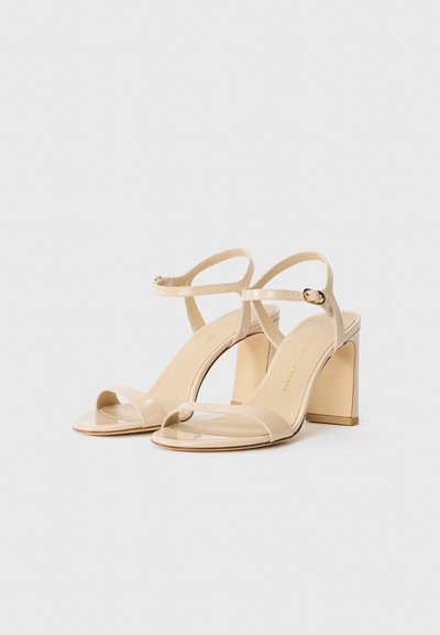 Beige patent leather avointen sandaalit, joissa on paksut korot ja nilkkahihnat, sekä kultainen solki valkoista taustaa vasten.