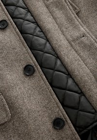 Tissu marron à motif chevrons avec une doublure noire matelassée. Dispose de boutons noirs et d'une coupe ajustée avec un col classique.