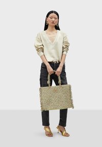 Borsa a tracolla intrecciata con fibre verdi e multicolori, con dettagli in frange e doppi manici. Il modello indossa una maglietta beige e jeans neri, con tacchi dorati.