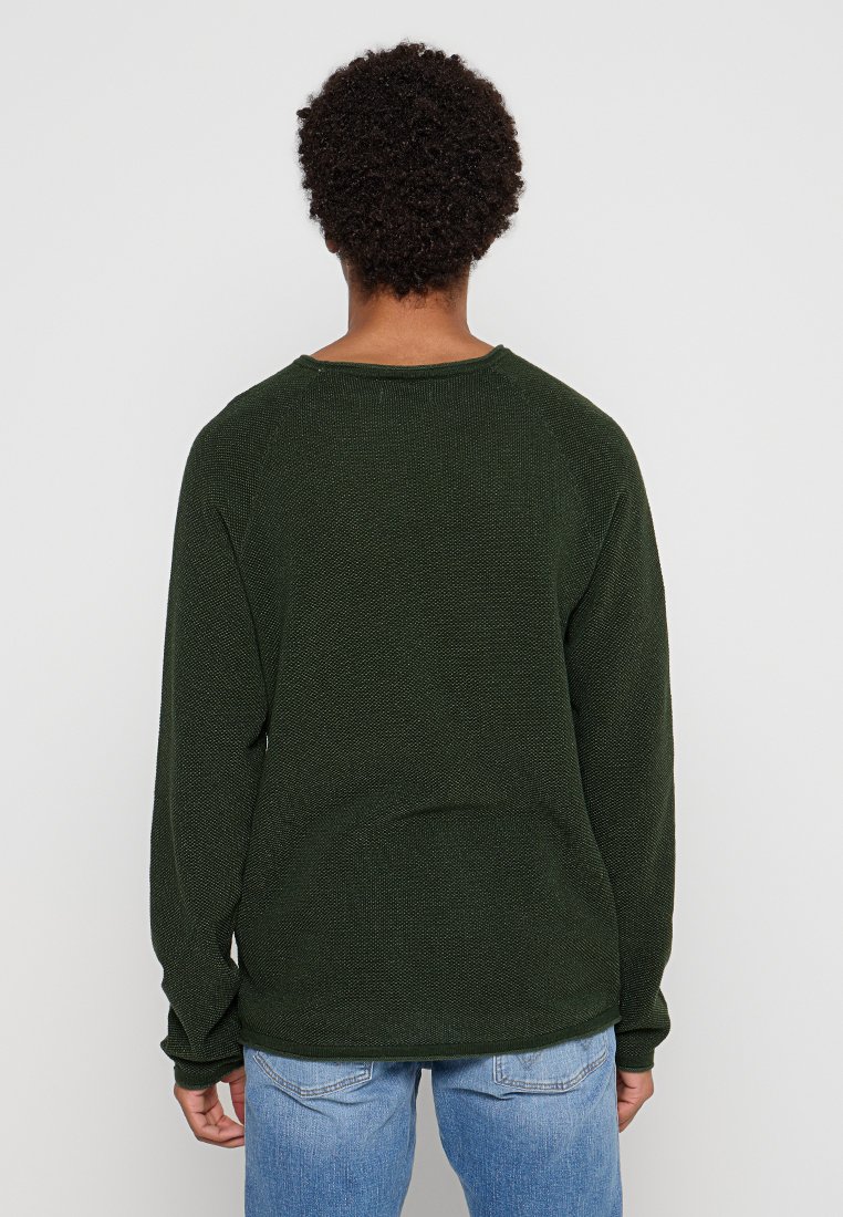 Donkergroene gebreide trui met een ronde halslijn, lange mouwen en een gematteerd oppervlak, gedragen over lichtblauwe denimjeans.