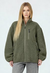 Veste en polaire vert olive avec une finition texturée, fermeture éclair à l'avant et deux poches latérales. Comprend un panneau lisse sur la poitrine et des détails zippés.
