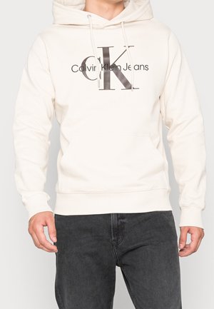 Jasna kremowa bluza z kapturem z kieszenią kangurzą z przodu, z odważnym szarym logo "CK" oraz tekstem "Calvin Klein Jeans" w czerni.