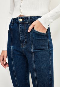 Džíny s vysokým pasem v tmavě modré barvě, viditelným prošíváním, předními kapsami a zlatým zapínáním na knoflík. Hladká textura s mírným pružením.