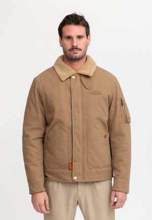 Man met kort donker haar en snor, gekleed in een lichtbruine jas met een schapenvachtkraag en beige broek, kijkt recht vooruit.
