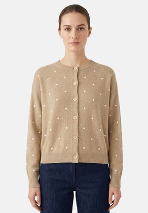 Donna che indossa un cardigan beige con bottoni con pois bianchi e jeans blu scuro, in piedi davanti a uno sfondo chiaro e uniforme.