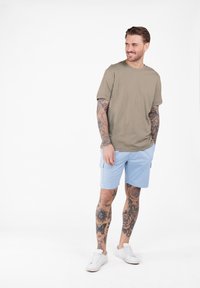 Jasnooliwkowa koszulka z krótkim rękawem zestawiona z jasnoniebieskimi szortami. Model ma widoczne tatuaże i nosi białe sneakersy.