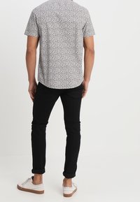 Camicia a maniche corte con motivo, sfondo chiaro con forme scure, abbinata a pantaloni neri aderenti e sneakers bianche con dettagli marroni.