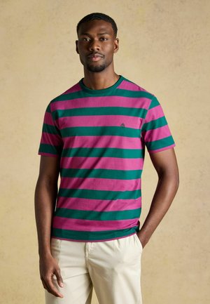 Homme debout, main dans la poche, portant une chemise à manches courtes rayée verte et rose et un pantalon beige, sur un fond beige uni.
