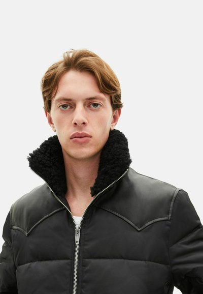 Schwarze Steppjacke mit durchgehendem Reißverschluss und strukturiertem schwarzem Kunstfellkragen. Glatte Oberfläche mit schrägen Schulternähten, figurbetontes Design.