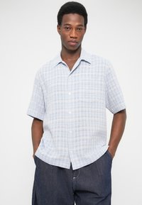 Νεαρός άνδρας με τα χέρια στις τσέπες, φοράει ένα ανοιχτό μπλε κοντομάνικο πουκάμισο με καρό σχέδιο και σκούρο μπλε παντελόνι, στέκεται μπροστά σε λευκό φόντο.