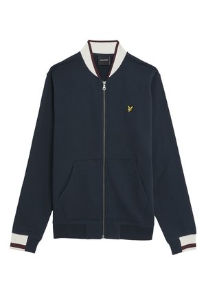 Marineblå zip-jacka i mjukt tyg, med en krämfärgad och mörkröd randig krage, två framfickor och ett litet gult emblem på bröstet.