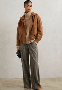 Jachetă din suede maro cu fermoar în față și guler, pulover bej cu gât înalt, pantaloni cu carouri și picioare largi cu dungi laterale, ținând o poșetă neagră.