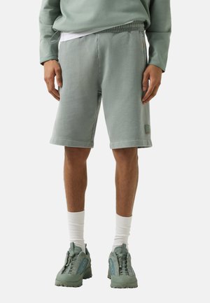 Mann trägt hellgrünes langärmeliges Hemd und passende mittelgroße Shorts mit weißen Socken und grünen Sportschuhen, steht mit entspannten Händen.