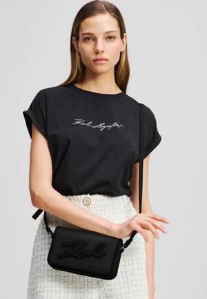 Sac à bandoulière en cuir noir avec logo "Karl" en relief, fermeture éclair, et sangle réglable, associé à un t-shirt noir à manches courtes et une jupe en tweed.